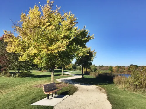 A walking path leads past a bench and autumn-colored trees around the pond