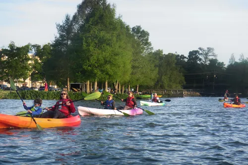 A group paddles colorful kayaks across University Lake, with the park in the background