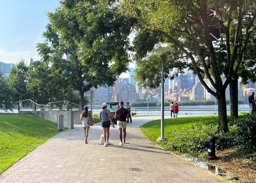 A group walks along the sunlit waterfront path with Manhattan visible across the East River