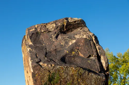 A large rock bearing ancient petroglyphs rises against a clear blue sky in the park